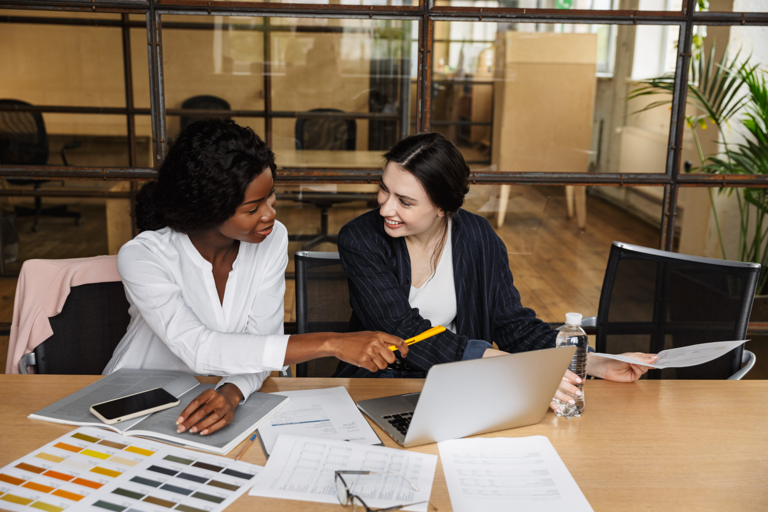 Two attractive smiling smart multiethnic women entrepreneurs working over new project in a modern office, sitting at the desk